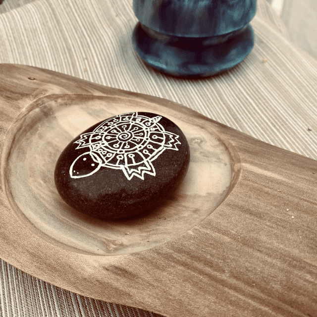 A painted stone with a mandala design resting on wooden platter, with a blue vase in the background